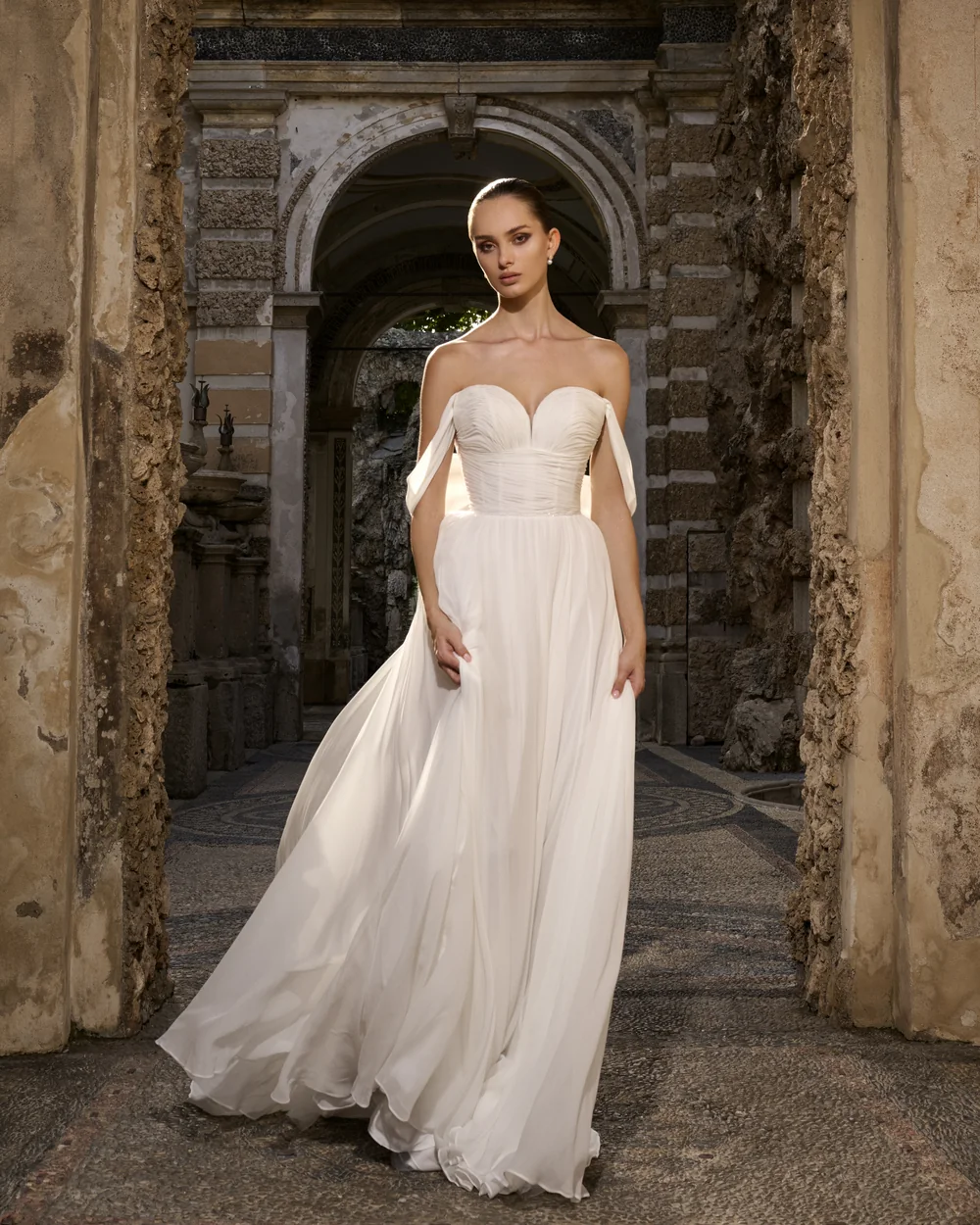 Sposa in abito di chiffon con corpetto drappeggiato, in piedi sotto un arco di pietra